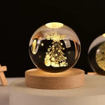 Illuminated glass crystal ball on a round wooden base, engraved with a glowing Christmas tree design with ornaments and small stars; warm light reflections on the sphere against a dark background, with another etched globe partially visible on the right.
