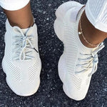 A pair of stylish white knitted athletic sneakers worn by a person standing on an asphalt surface. The shoes feature a breathable mesh design, thick cushioned soles, and gray laces. The person is also wearing white pants and a gold anklet.