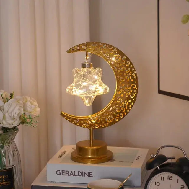 A decorative lamp featuring a golden crescent moon with intricate cut-out patterns. The moon cradles a star-shaped light fixture with glowing fairy lights inside. The lamp is placed on a table with a book titled "GERALDINE" and other objects nearby.