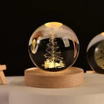 Illuminated glass crystal ball on a round wooden base, engraved with a glowing Christmas tree design with ornaments and small stars; warm light reflections on the sphere against a dark background, with another etched globe partially visible on the right.