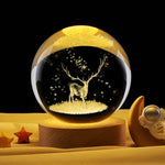 Glowing crystal ball lamp on a wooden base, featuring an illuminated deer silhouette with antlers and floating star-like sparkles, set on a yellow surface with small star decor and a moon-and-astronaut figurine nearby.