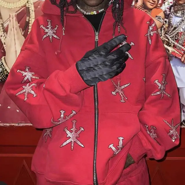 A red zip-up hoodie with a relaxed streetwear fit, featuring an all-over silver cross-style embroidered graphic pattern on the sleeves and body. It has a drawstring hood, front zipper closure, ribbed cuffs, and front pockets. Shown on a person wearing black gloves, the hoodie has a bold edgy look with standout decorative detailing.