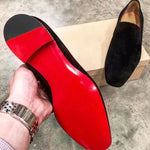A pair of elegant burgundy leather loafers with a glossy finish. The shoes feature gold-toned metal horsebit detailing across the uppers and are displayed with wooden shoe trees inserted. They have a sleek, elongated silhouette with subtle stitching along the toe cap. The loafers are placed on a dark surface, partially framed by a wooden picture frame, emphasizing their classic and sophisticated style.