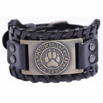 A black leather wristband with a braided design and a bronze-colored circular clasp featuring a paw print and runic inscriptions.
