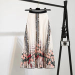 Pleated midi skirt with a gradient design, transitioning from dark black at the waistband to a soft blush pink at the hem, hanging on a black clothing rack next to a vintage-style camera.
