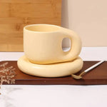 Cream-colored ceramic mug with a rounded, cutout-style handle, sitting on a matching donut-shaped saucer; a small spoon rests on a wooden tray beside it.