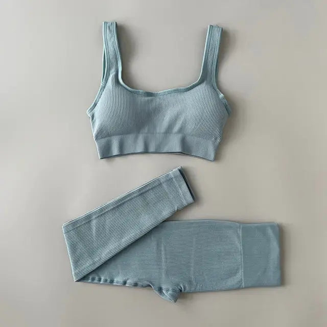 Black ribbed sports bra and matching high-waisted leggings set laid flat on a neutral gray background. The outfit is designed for activewear or athleisure, featuring a scoop neckline and wide straps on the bra.