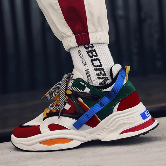 Close-up of a multicolor chunky sneaker (white, red, green, and yellow) with patterned laces and a blue side strap, worn with white “SPORT” socks and jogger pants.