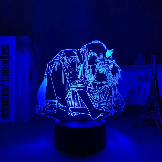 A blue LED acrylic night light displaying an anime-style couple in an intimate pose, glowing against a dark room. The lamp sits on a round black base, with books and a small Eiffel Tower decoration blurred in the background.