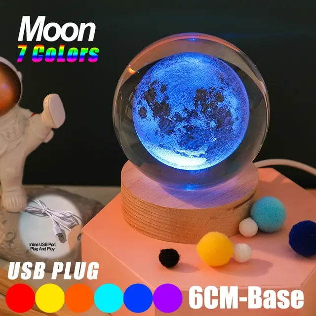 Glowing 3D moon crystal ball lamp on a wooden base, lit in blue, with text reading “Moon 7 Colors,” “USB Plug,” and “6CM-Base,” plus colored pom-poms on a tabletop.
