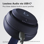 Close-up promotional image of a dark blue Beats over-ear headphone earcup with a USB-C cable plugged in, with the text “Lossless Audio via USB-C” and “Plus three distinct built-in sound profiles” on a light gray background.