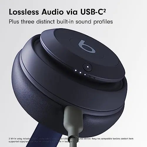 Close-up promotional image of a dark blue Beats over-ear headphone earcup with a USB-C cable plugged in, with the text “Lossless Audio via USB-C” and “Plus three distinct built-in sound profiles” on a light gray background.