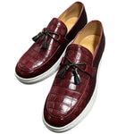 A pair of elegant burgundy leather loafers with a glossy finish. The shoes feature gold-toned metal horsebit detailing across the uppers and are displayed with wooden shoe trees inserted. They have a sleek, elongated silhouette with subtle stitching along the toe cap. The loafers are placed on a dark surface, partially framed by a wooden picture frame, emphasizing their classic and sophisticated style.