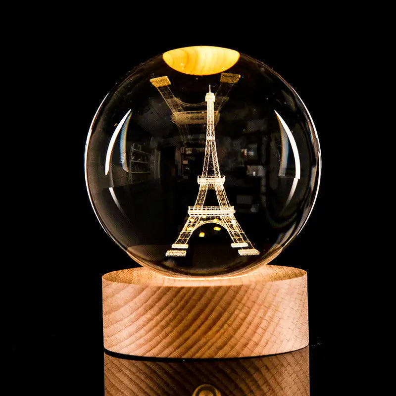Crystal ball lamp with a laser-etched Eiffel Tower inside, glowing warm light on a wooden base against a black background.