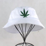 White bucket hat with an embroidered green leaf emblem on the front, displayed on a metal stand against a light background.