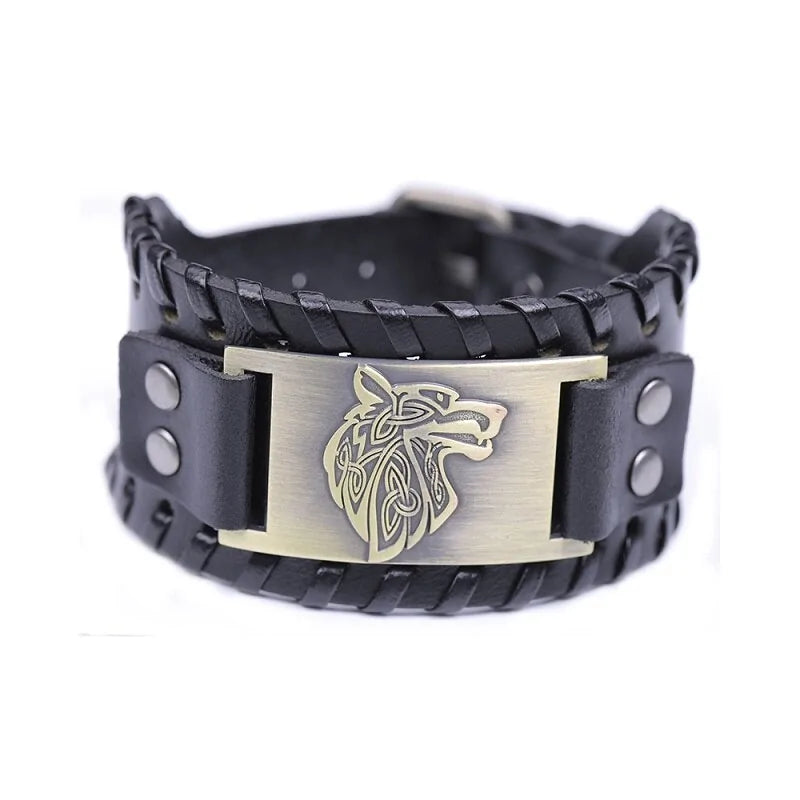 A chunky black leather wrist cuff is shown against a white background. The bracelet has braided edging along the top and bottom and is fastened with riveted leather tabs on both sides. In the center is a brushed metal plate with a gold-toned, Celtic-style wolf head emblem. The overall style looks rugged and masculine, with layered leather and metal detailing.