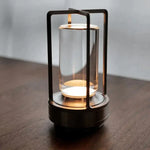 Modern tabletop lantern with a black frame and clear cylindrical glass shade, emitting a warm light on a wooden surface.