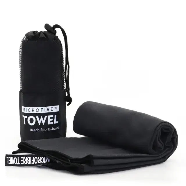 Black microfiber towel shown folded and rolled on a white background, beside a black drawstring mesh carry bag labeled “MICROFIBER TOWEL” with “Beach·Sports·Travel” underneath.
