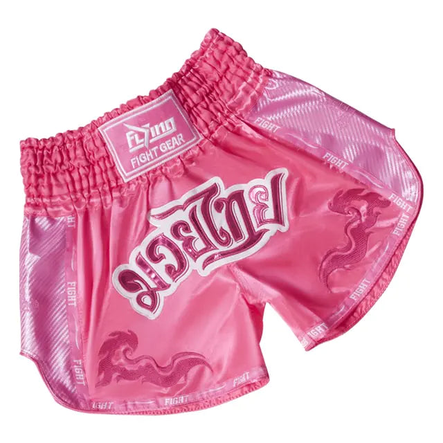 Pink satin Muay Thai boxing shorts with an elastic waistband, white Thai-style lettering on the front, and “FIGHT” trim along the edges, on a white background.