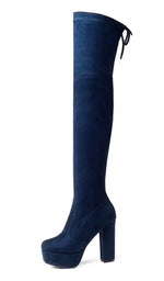 A woman wearing black high-heeled, over-the-knee boots with an open lattice cutout design running up the back, revealing skin between the horizontal straps. She stands indoors on a light wooden floor near a white ornate console table.