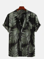 A studio product image of a black short-sleeve button-up shirt on a hanger, featuring an all-over green fern/leaf print, a spread collar, rolled sleeve cuffs, and white buttons, photographed against a light background.