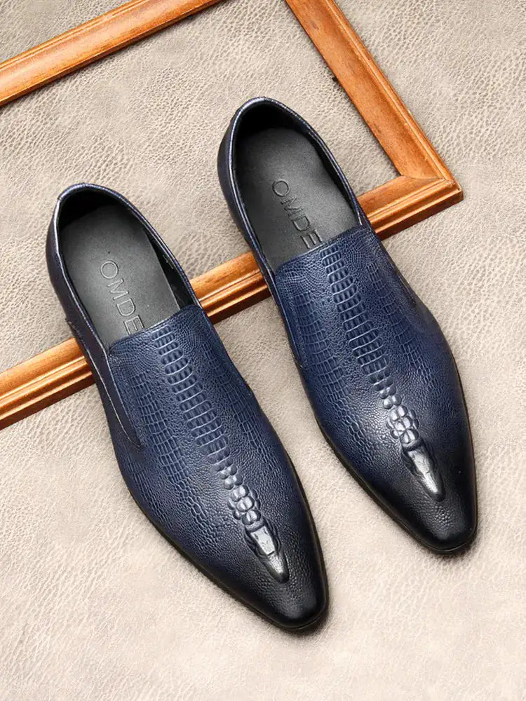 A pair of elegant burgundy leather loafers with a glossy finish. The shoes feature gold-toned metal horsebit detailing across the uppers and are displayed with wooden shoe trees inserted. They have a sleek, elongated silhouette with subtle stitching along the toe cap. The loafers are placed on a dark surface, partially framed by a wooden picture frame, emphasizing their classic and sophisticated style.