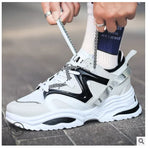 A white chunky sneaker with layered panel detailing, black side overlays, patterned laces, and a thick sculpted sole. The shoe is shown being laced up, paired with white socks and set against an outdoor pavement background.
