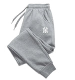 A pair of light gray sweatpants with an elastic waistband and drawstrings, laid flat against a white background. The sweatpants feature ribbed cuffs at the ankles and side pockets. A black embroidered logo, resembling the New York Yankees emblem, is visible on the upper left thigh. The fabric appears to be soft, suggesting a comfortable and relaxed fit.