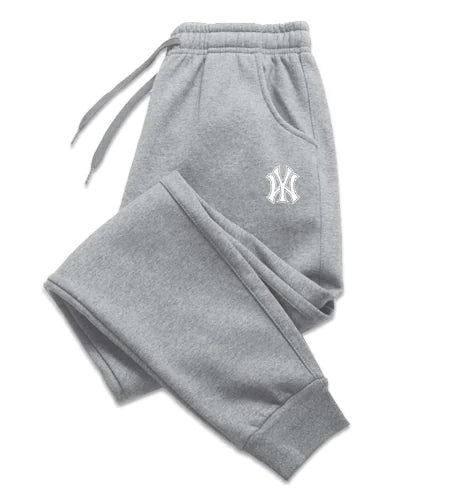 A pair of light gray sweatpants with an elastic waistband and drawstrings, laid flat against a white background. The sweatpants feature ribbed cuffs at the ankles and side pockets. A black embroidered logo, resembling the New York Yankees emblem, is visible on the upper left thigh. The fabric appears to be soft, suggesting a comfortable and relaxed fit.