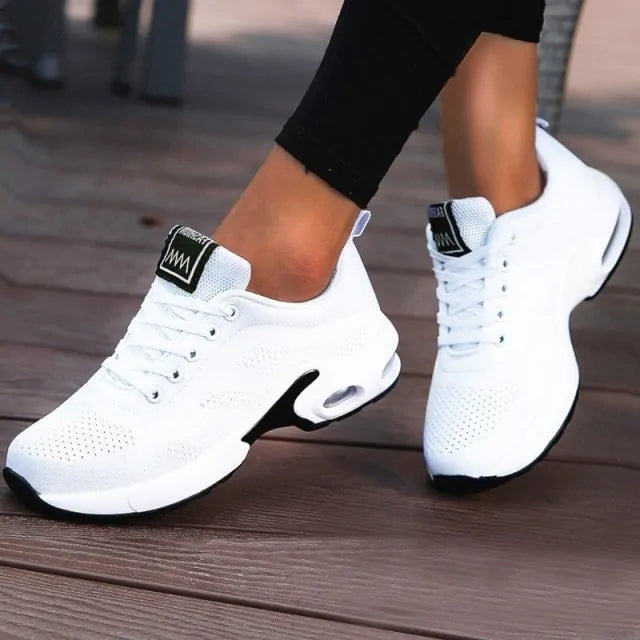 A black and dark gray athletic sneaker with a breathable knit upper, accented by bold orange wave-like stripes on the sides and orange detailing on the midsole. The shoe features a thick, textured sole and black-and-white patterned laces, designed for both style and performance.