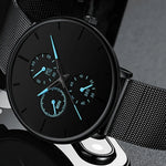 Close-up of a black minimalist wristwatch with a mesh metal strap, featuring three small subdials and blue-accented hands and markers on a matte black face.
