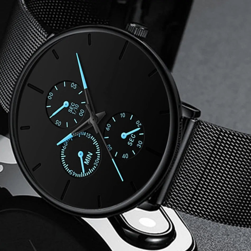 Close-up of a black minimalist wristwatch with a mesh metal strap, featuring three small subdials and blue-accented hands and markers on a matte black face.