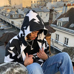 Person sitting on a rooftop wearing a black-and-white star-pattern hoodie, layered necklaces, and blue jeans, with city buildings and chimneys in the background.