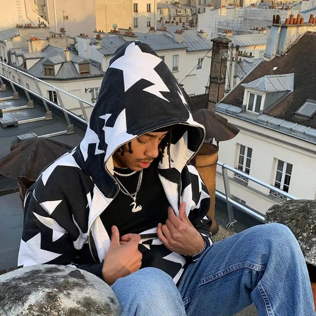 Person sitting on a rooftop wearing a black-and-white star-pattern hoodie, layered necklaces, and blue jeans, with city buildings and chimneys in the background.