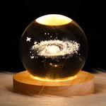 A glowing crystal ball lamp on a round wooden base, lit warm yellow and etched with a spiral galaxy and small stars against a black background.