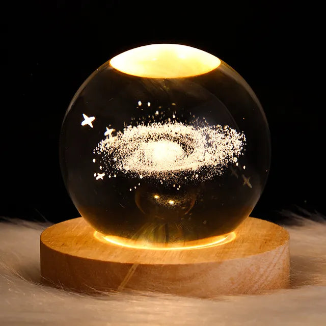A glowing crystal ball lamp on a round wooden base, lit warm yellow and etched with a spiral galaxy and small stars against a black background.