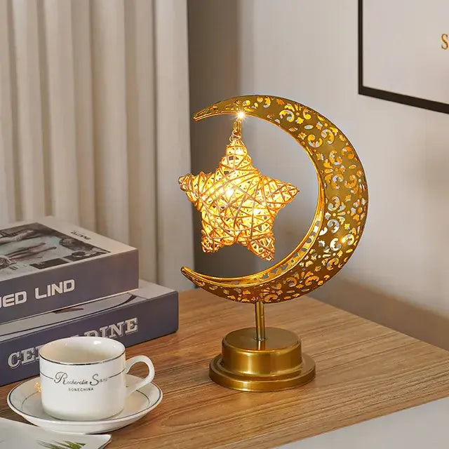 Gold crescent moon tabletop lamp with a glowing hanging star inside, sitting on a wooden table beside books and a teacup.