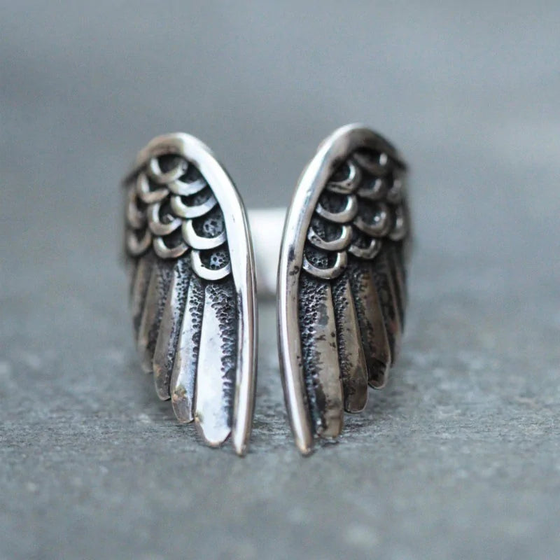 Close-up of an oxidized silver ring shaped like a pair of angel wings, with layered feather details on a gray surface.