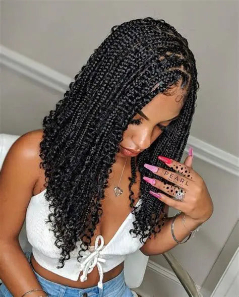 Woman with shoulder-length box braids, styled in loose curls, wearing a white cropped top and light blue jeans. She is also showing off long pink manicured nails.
