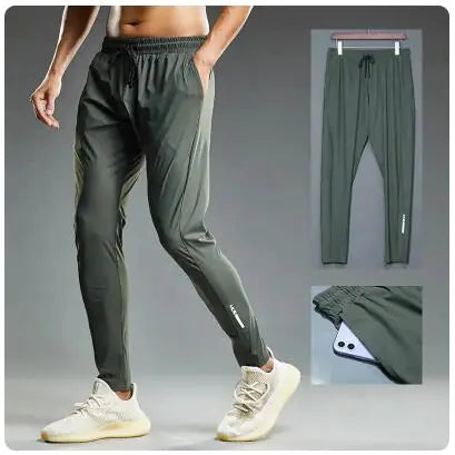 A man models a pair of olive-green athletic joggers with a slim fit, elastic waistband, and drawstring for an adjustable fit. The joggers feature a subtle reflective design on the lower leg, adding a sporty touch. The image also includes two smaller insets: one showing the joggers hanging on a hanger, and another highlighting a zippered pocket with a smartphone partially tucked inside, emphasizing convenience and functionality. The model pairs the joggers with white sneakers, making them suitable for casual