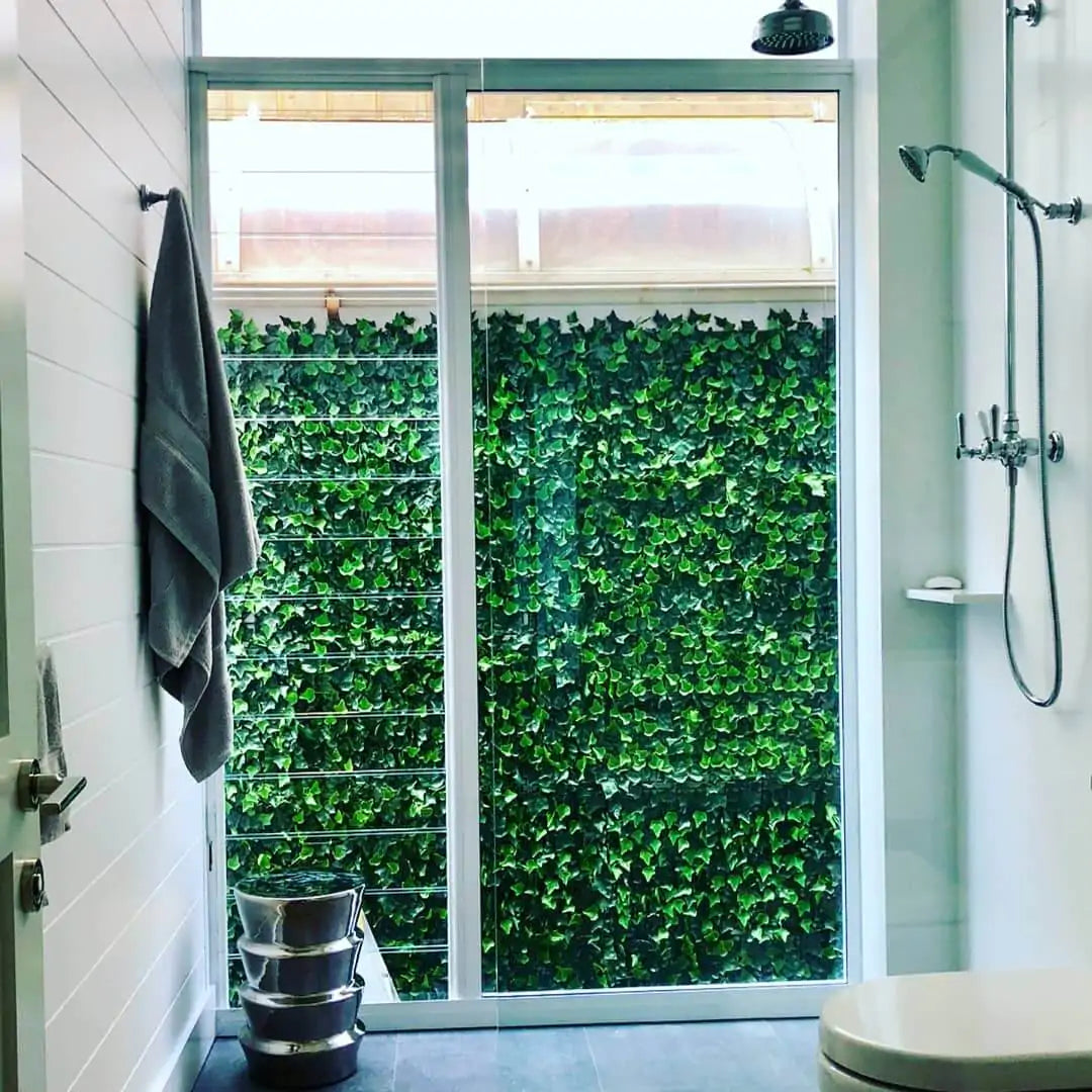 Bright modern bathroom shower with a large glass sliding door opening to a lush green ivy-covered wall outside, featuring a rainfall showerhead, handheld shower, and a towel hanging on the wall.