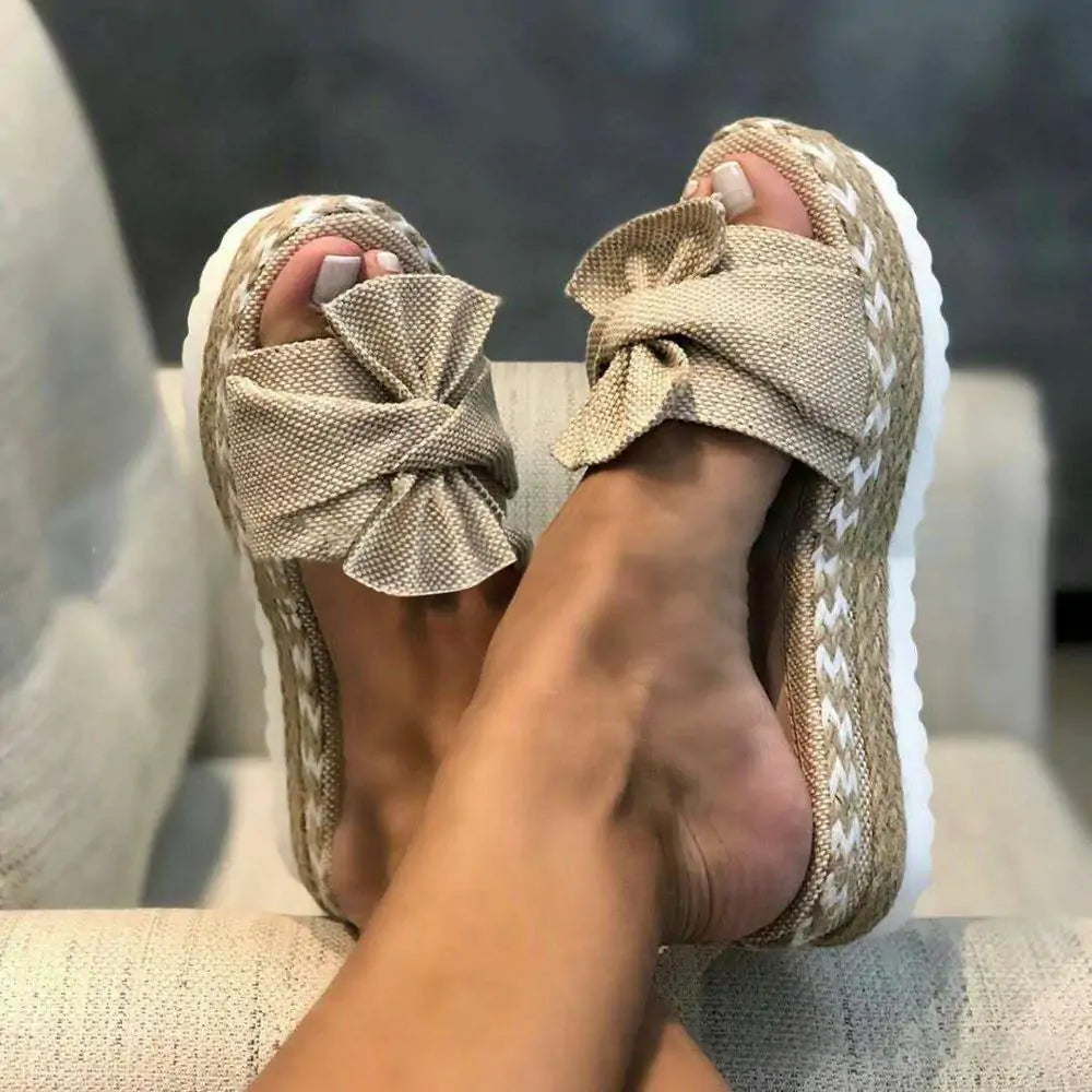 A pair of beige open-toe platform slide sandals with oversized bow details across the front. The sandals have woven espadrille-style sides with white braided trim and thick white soles, shown worn while resting on a light upholstered chair.