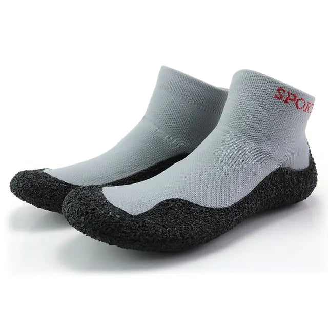 A pair of minimalist sock-style water shoes in a green and black knit pattern, featuring black ankle cuffs and textured black rubber soles that wrap around the lower sides for added grip.