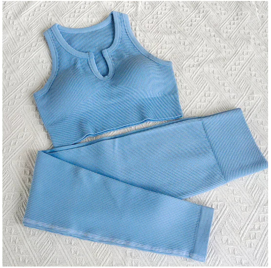 Black ribbed sports bra and matching high-waisted leggings set laid flat on a neutral gray background. The outfit is designed for activewear or athleisure, featuring a scoop neckline and wide straps on the bra.