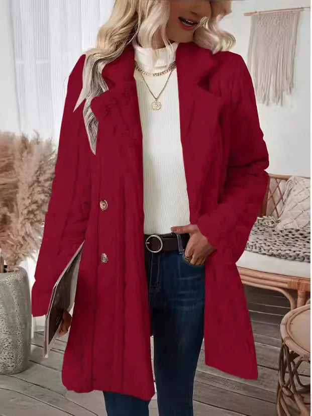 Stylish woman wearing a long red buttoned coat over a white ribbed turtleneck sweater, paired with dark blue jeans and accessorized with layered gold necklaces and a black belt. She is holding a magazine and standing in a cozy, modern room with neutral decor.