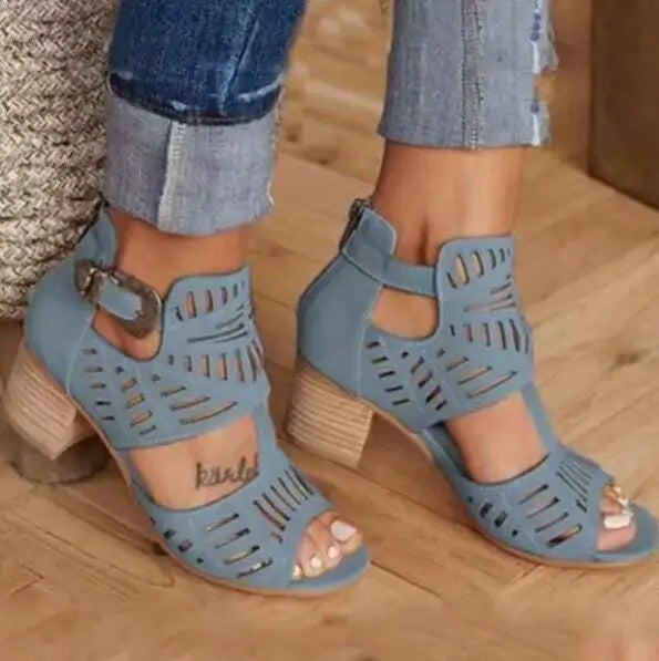 A close-up view of a person wearing stylish beige cut-out heeled sandals with ankle straps and decorative perforations, paired with cuffed blue denim jeans on a wooden floor.