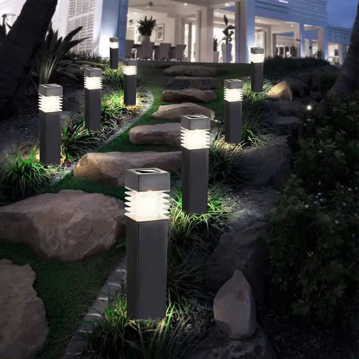 Nighttime garden pathway with large stepping stones and landscaping, illuminated by a row of modern black bollard lights casting bright white light toward a house in the background.
