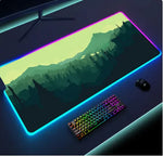 Large RGB-lit gaming mouse pad with a mountain-and-forest design on a desk, shown with a rainbow-backlit keyboard and a mouse beside it.