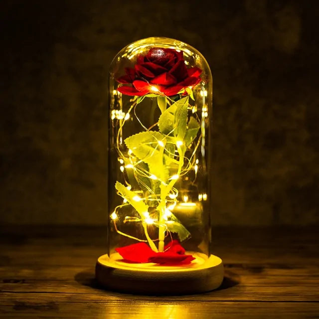 Glowing decorative rose lamp: a red rose inside a clear glass dome wrapped with warm white fairy lights on a round base, set against a dark background.