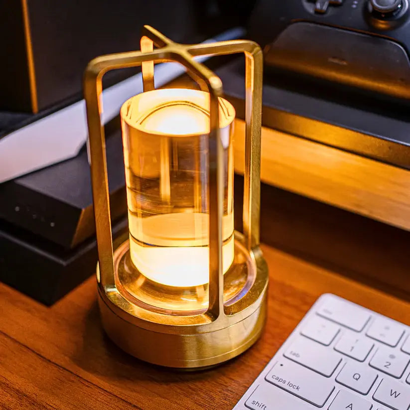 Warm-lit brass table lamp with a cylindrical glass shade inside a geometric metal frame, sitting on a wooden desk next to a keyboard and other desk items.